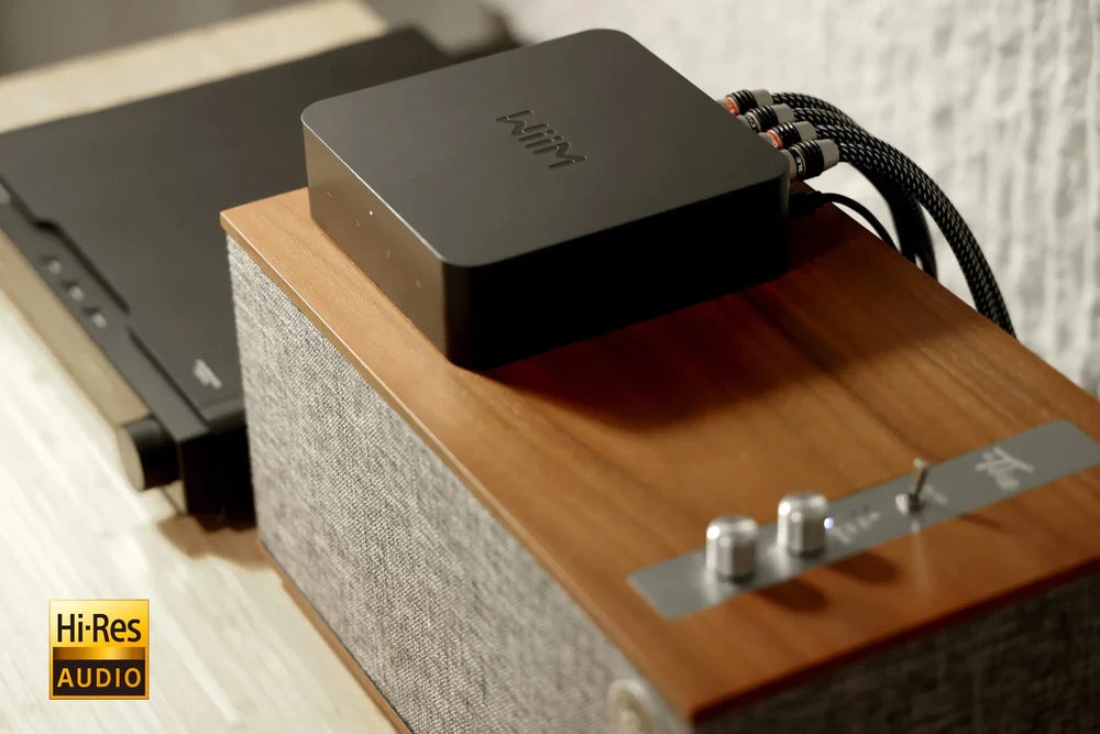 GPT Das Bild zeigt einen schwarzen WiiM Pro Streaming-Adapter, der auf einem hölzernen Lautsprecher platziert ist. Der Lautsprecher ist teilweise von einem grauen Stoff bedeckt, und am unteren Rand des Bildes ist das gelbe Hi-Res AUDIO-Logo zu sehen.