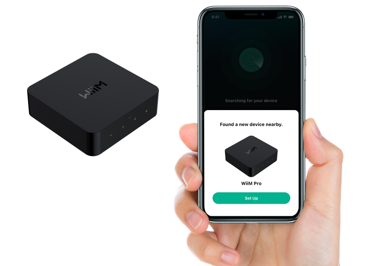 Das Bild zeigt eine Hand, die ein Smartphone hält, auf dessen Bildschirm die Einrichtung eines neuen Geräts, des "WiiM Pro", angezeigt wird. Neben dem Smartphone schwebt das Bild eines schwarzen, quadratischen WiiM Pro-Geräts