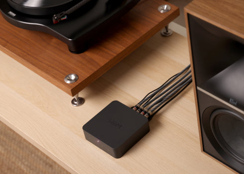 Das Bild zeigt einen schwarzen WiiM Pro Streaming-Adapter, der auf einem Holzregal liegt, mit mehreren angeschlossenen Audiokabeln, die aus ihm herausführen. Im Hintergrund ist ein Teil eines Plattenspielers und der Rand eines Standlautsprechers zu sehen.