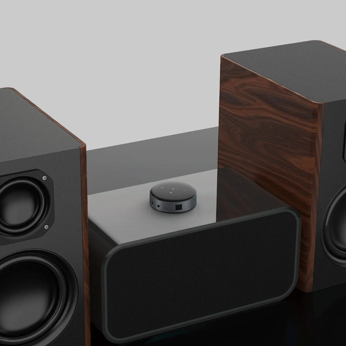Das Bild zeigt eine Nahaufnahme eines WiiM Mini, platziert auf einem grauen Regal, flankiert von zwei großen Standlautsprechern mit sichtbaren Treibern und einer Holzverkleidung. Der WiiM Mini ist ein kleines rundes Gerät in Schwarz mit berührungsempfindlichen Tasten auf der Oberseite und verschiedenen Anschlüssen an der Seite. Das Bild hat einen neutralen Hintergrund, der die Fokus auf das Audio-Gerät legt.