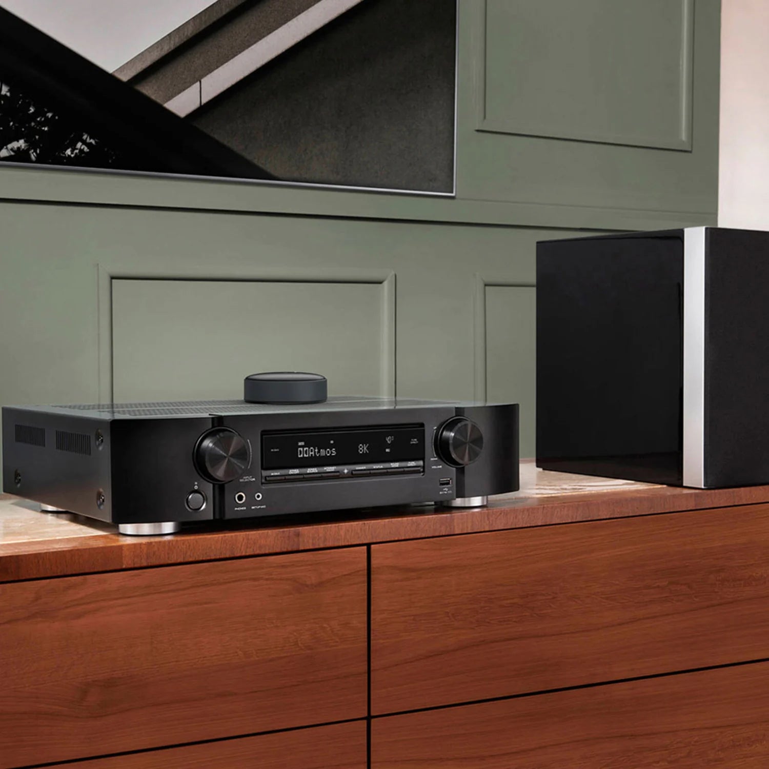 Das Bild zeigt ein modernes Wohnzimmer-Entertainment-Setup mit einem schwarzen AV-Receiver, auf dem ein WiiM Mini liegt, ein kompaktes rundes Streaming-Gerät. Rechts davon steht ein großer, zweifarbiger Standlautsprecher. Die Geräte befinden sich auf einer Holzkonsole mit sichtbarer Maserung, und im Hintergrund ist eine Wand in einem tiefen Grünton mit Wandpaneelen zu sehen. 